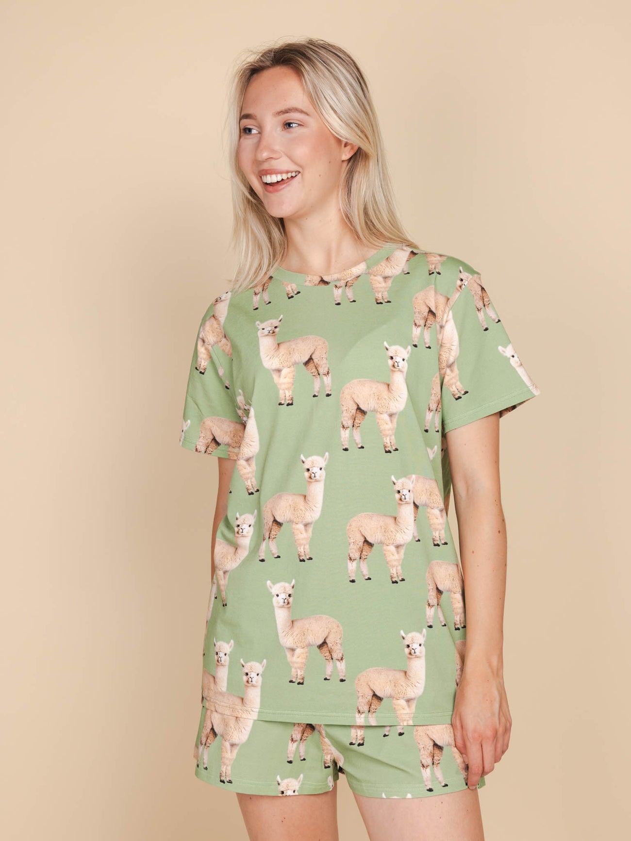 SNURK Alpaca Meadow T-shirt Unisex met alpacaprint gedragen door een vrouw op een beige achtergrond.