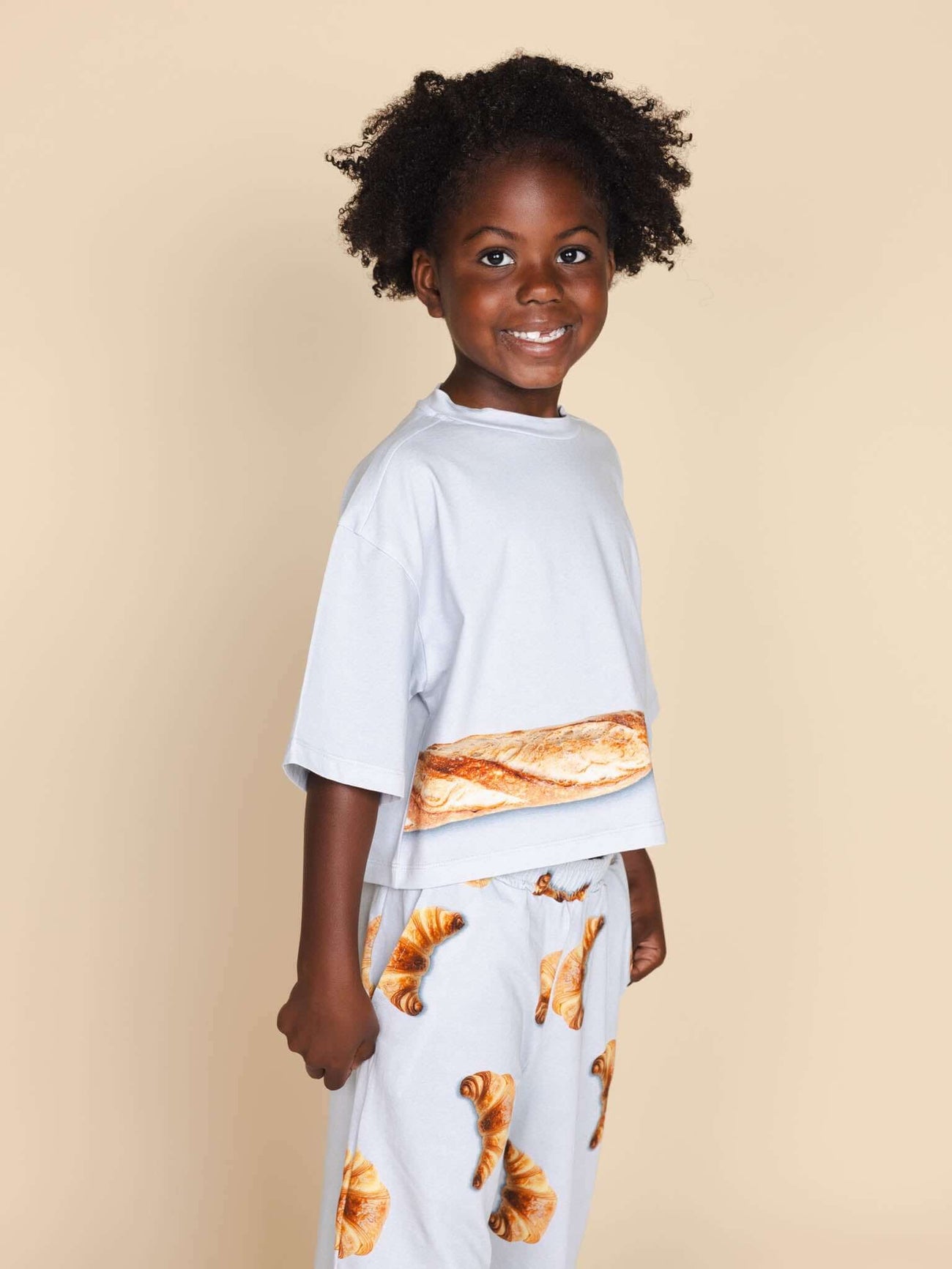 SNURK Baguette Cropped T-Shirt Kids met een vrolijk kind in een pyjama met croissantmotief.