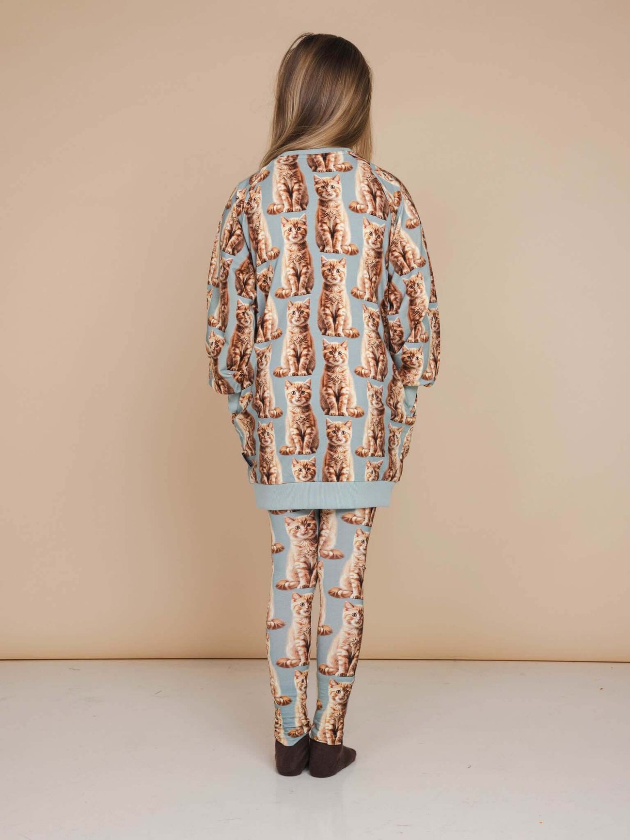 A girl wearing the SNURK Bowie Sweater Dress Kids with an all-over print of playful orange cats.