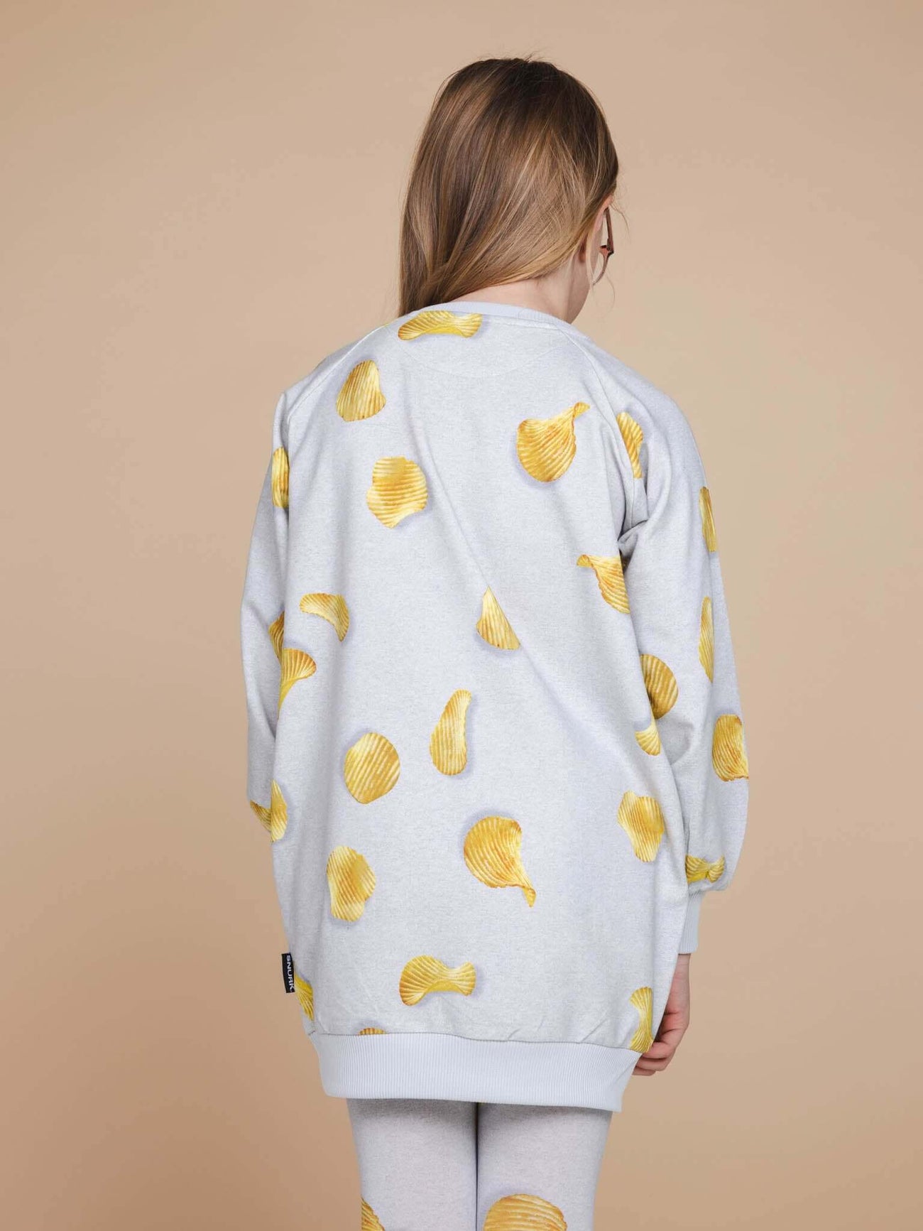 A child wearing the SNURK Chips Sweater Dress Kids featuring a playful all-over print of chips on a light grey background.