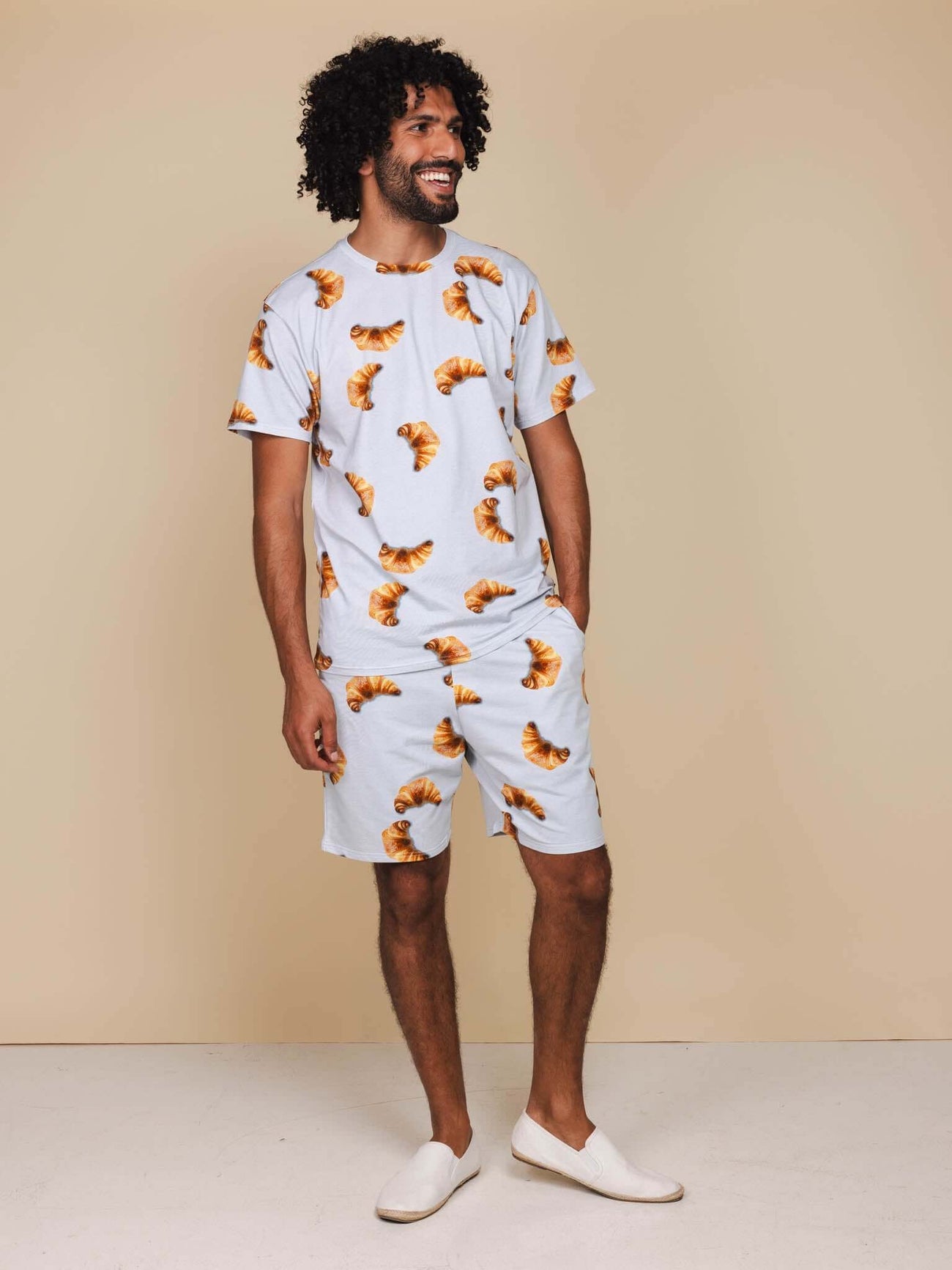 Man in SNURK Croissants Shorts Heren met afgebeelde croissantjes op een lichtblauwe achtergrond.