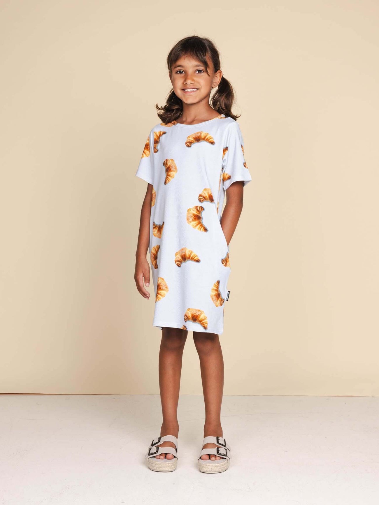 SNURK Croissants T-shirt Dress Kids gedragen door een meisje met een vrolijke uitstraling, gekleed in een jurkje met croissants.