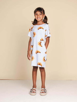 SNURK Croissants T-shirt Dress Kids gedragen door een meisje met een vrolijke uitstraling, gekleed in een jurkje met croissants.