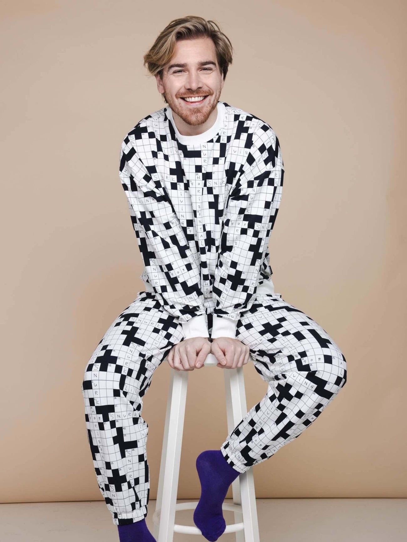 SNURK Crossword Sweater Heren model zittend op een kruk in zwart-witte kruiswoordprint outfit met paarse sokken.