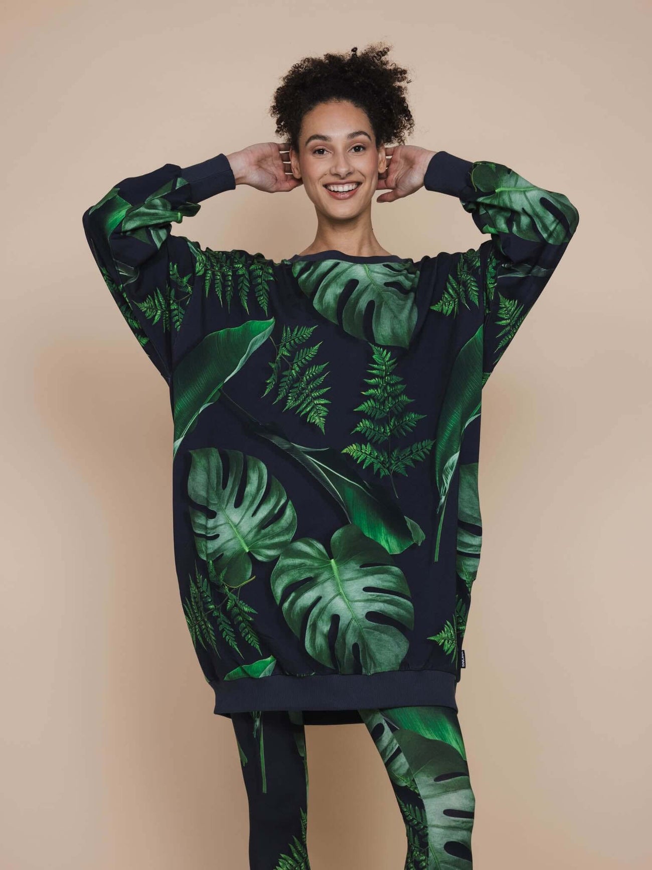 Model toont de SNURK Dark Forest Legging Dames met grote bladeren op een antracietgrijze achtergrond.