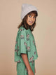 Meisje met een beanie, gekleed in het SNURK Donkeys Cropped T-Shirt Kids met schattige baby-ezels op een groene achtergrond.