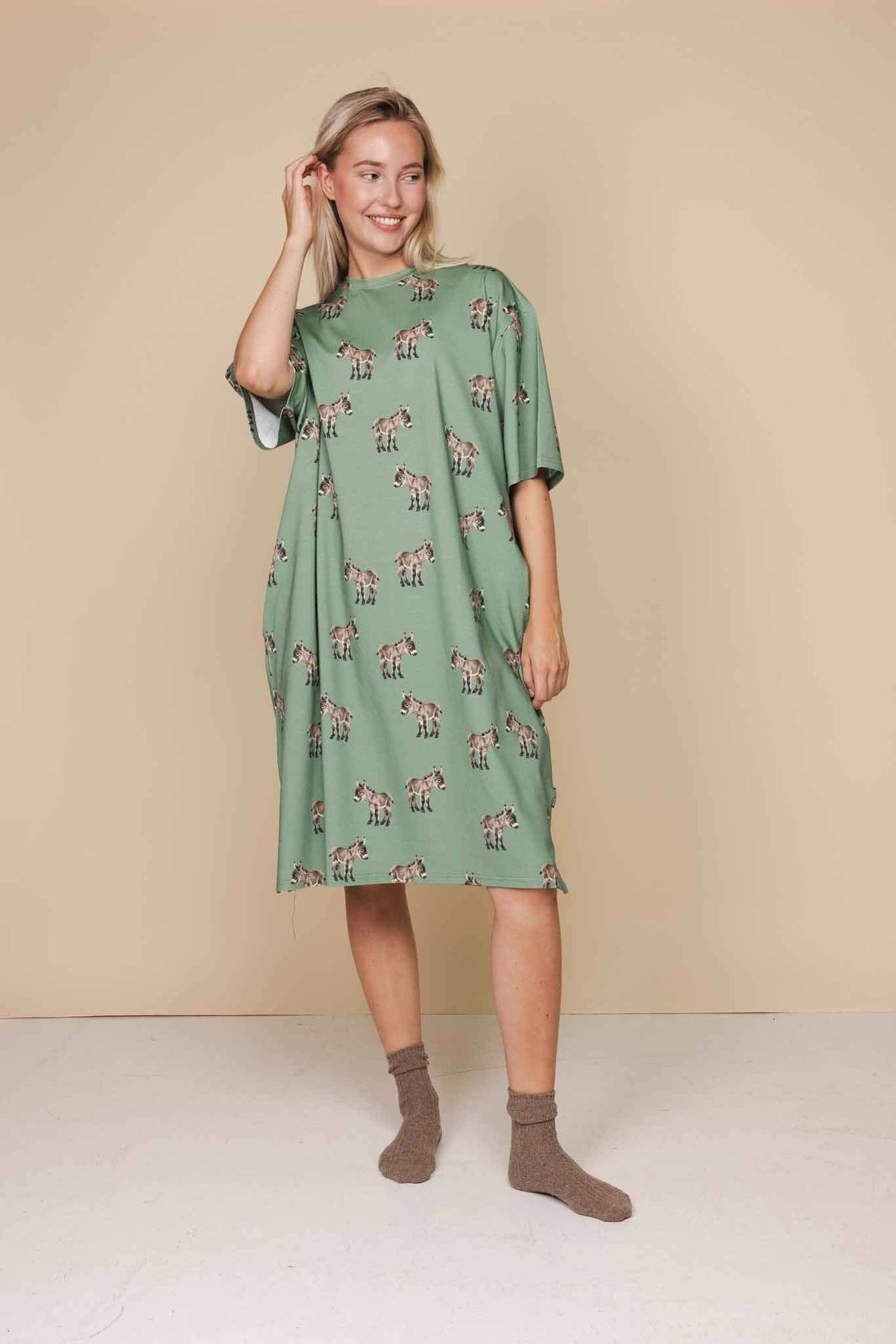 Model draagt SNURK Donkeys Relaxed Tee Dress Dames met schattige baby-ezels op een groene achtergrond.