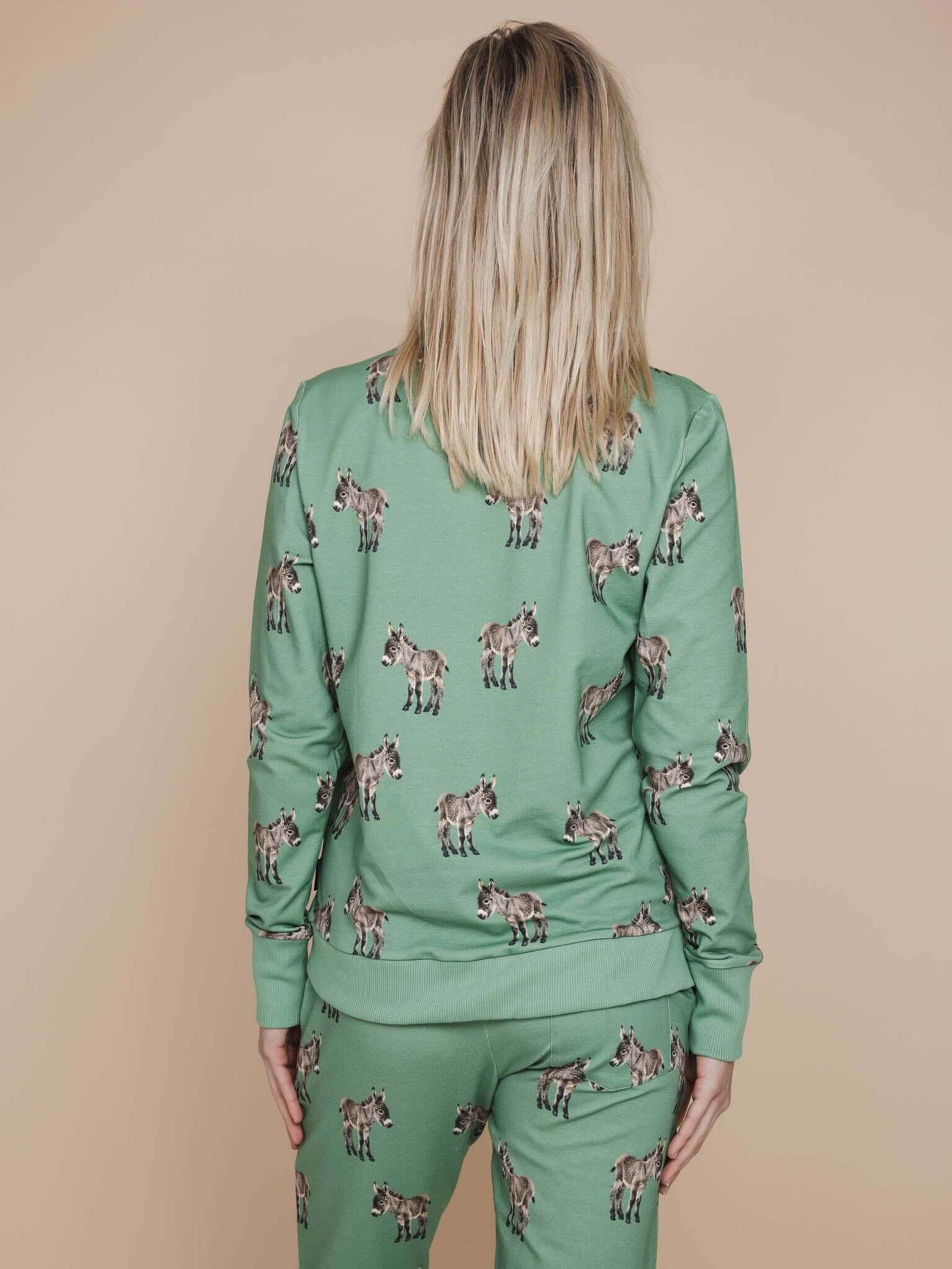 Een vrouw draagt de SNURK Donkeys Sweater Dames met schattige baby-ezels op een zachte groene achtergrond.