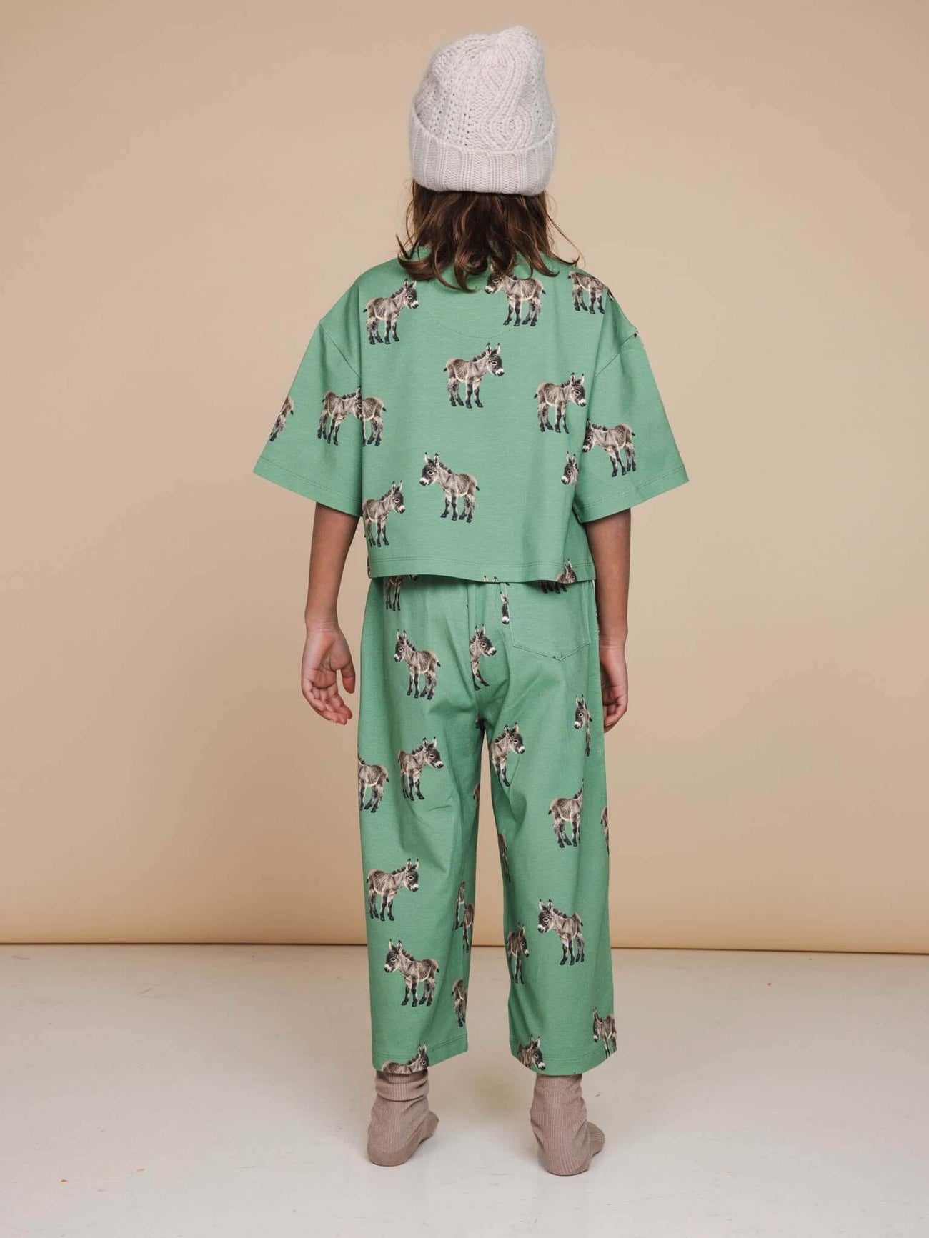 Achteraanzicht van een kind in een SNURK Donkeys Wijde broek Kids met schattige baby-ezels op een zachtgroene achtergrond.
