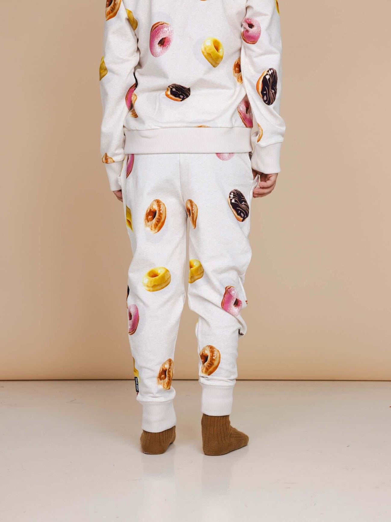 Kind draagt SNURK Donuts Broek Kids met kleurrijke donutprint op een beige achtergrond.