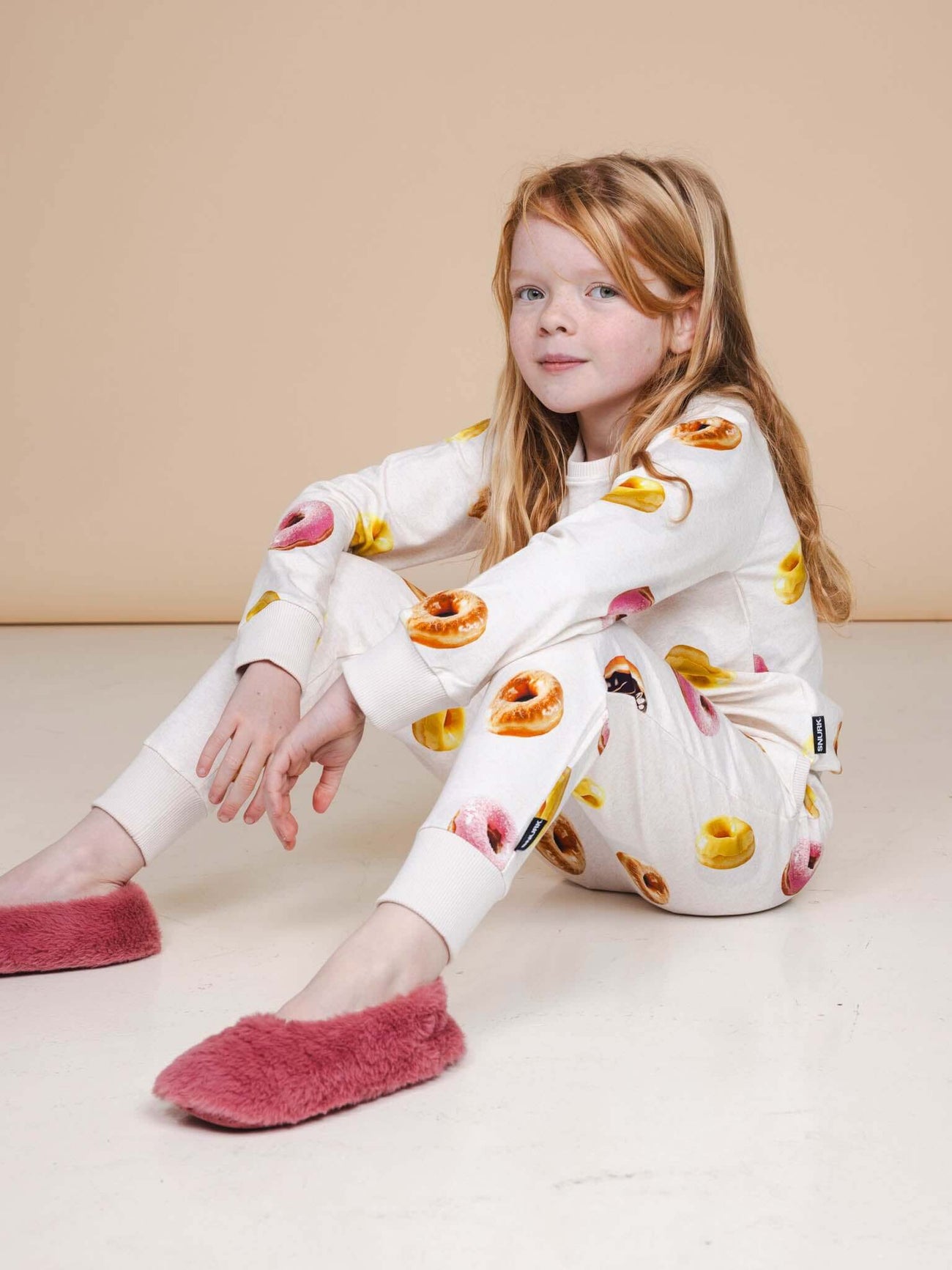 Meisje in SNURK Donuts Broek Kids met kleurrijke donutprint, zittend op de vloer in zachte pantoffels.