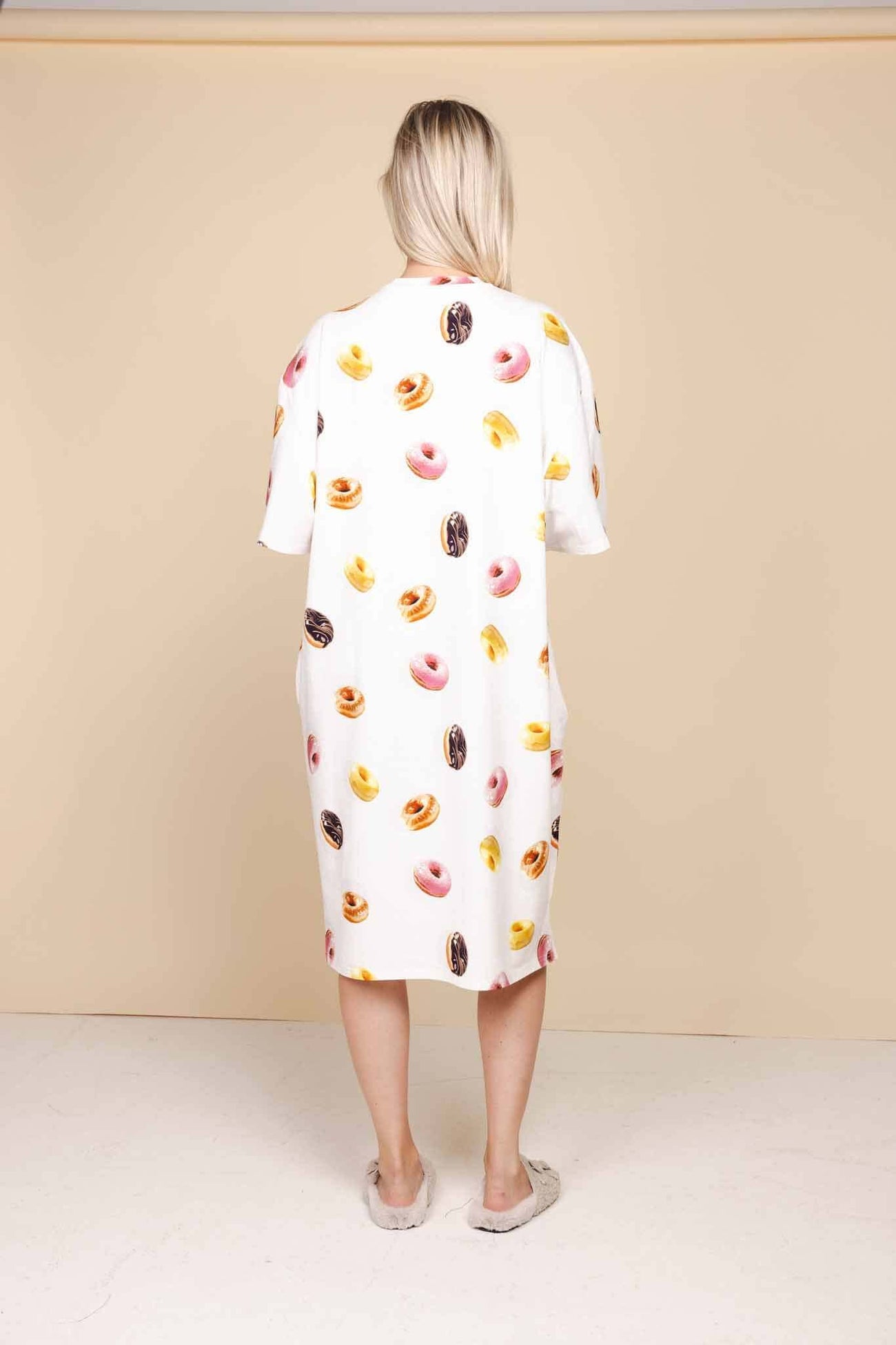 A woman wearing the SNURK Donuts Relaxed Tee Dress Dames, featuring a colorful donut print on a soft beige background.