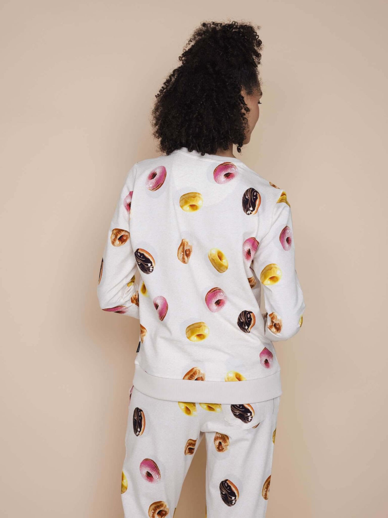 Vrouwelijke model draagt de SNURK Donuts Sweater Dames met kleurrijke donutprint op een beige achtergrond.