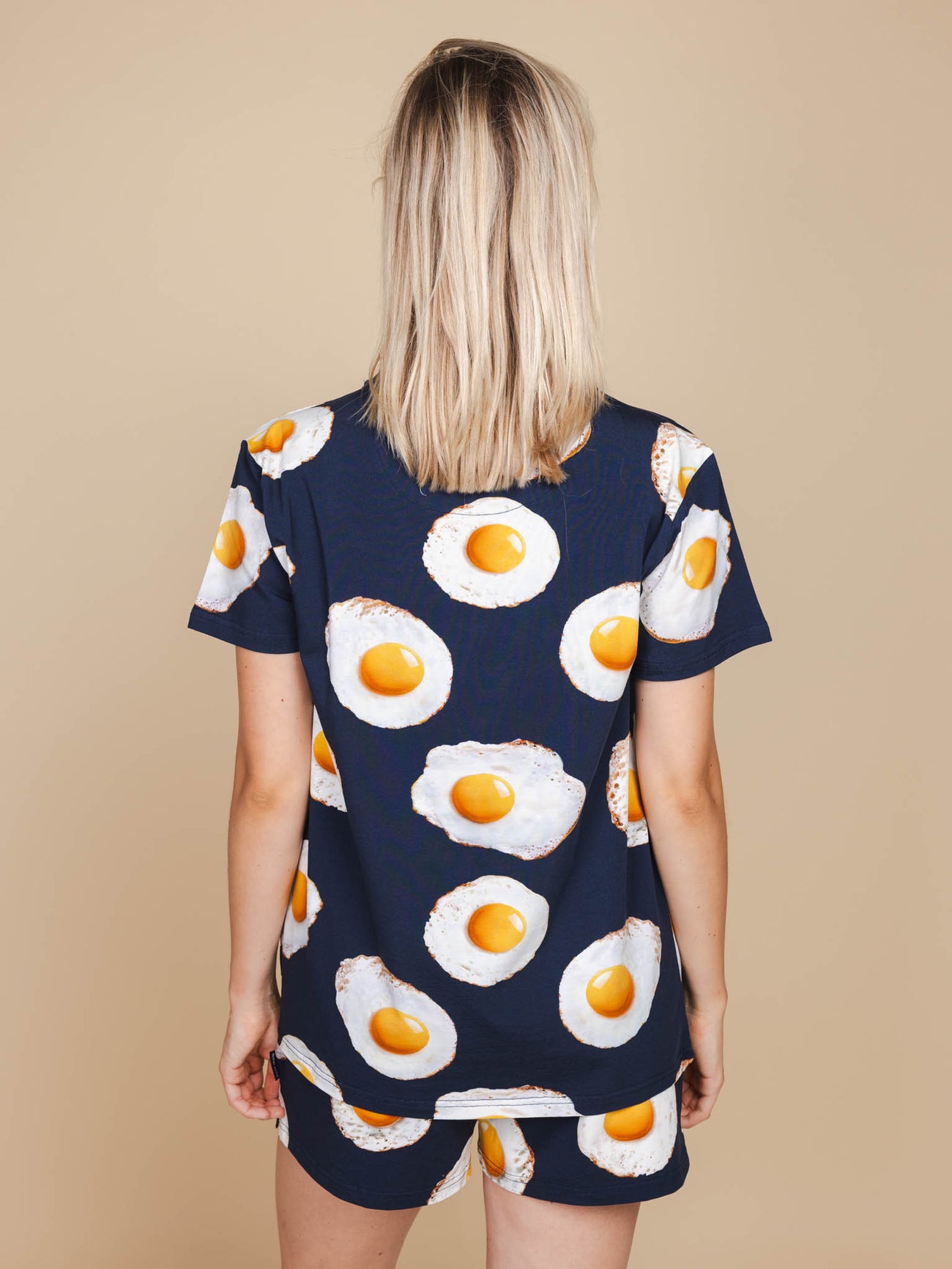 Achterkant van een vrouw die het SNURK Eggs Navy T-shirt Unisex draagt met spiegeleitjes op een blauwe achtergrond.