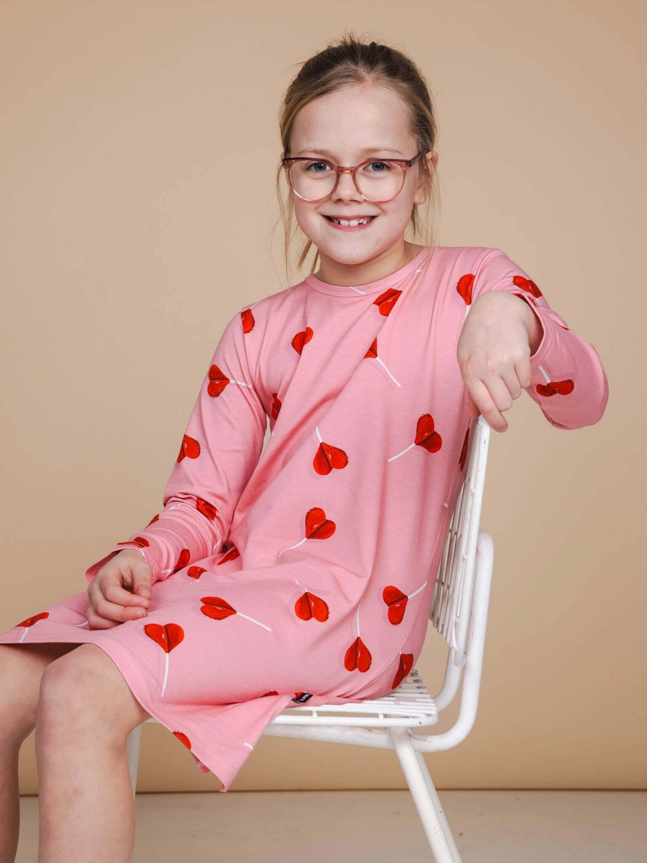 Meisje in SNURK Lollipop Love Long Sleeve Dress Kids met hartjeslollies op roze, zittend op een witte stoel.