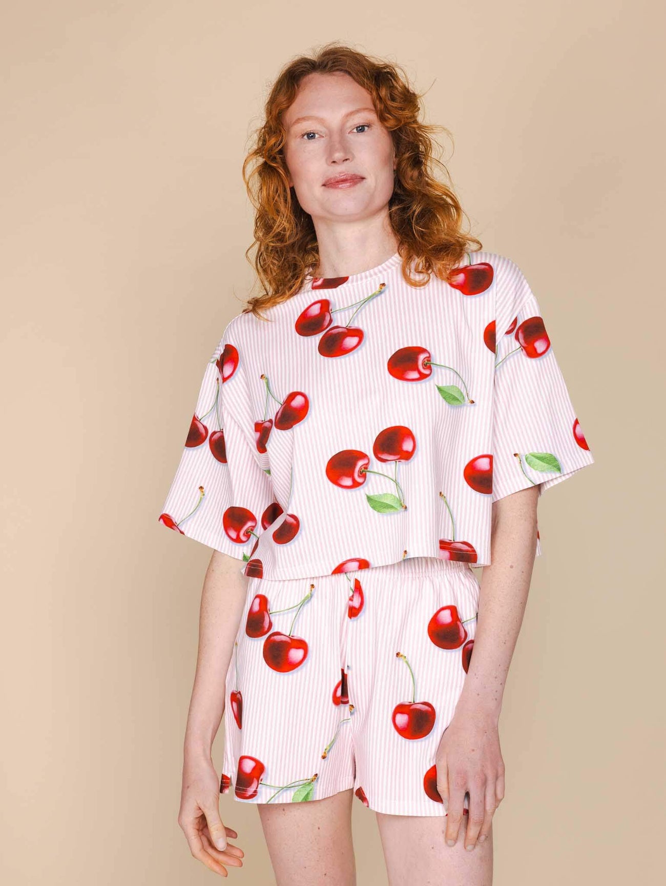 Model in SNURK Mon Cherry Shorts Dames met rode kersenprint op een beige achtergrond.