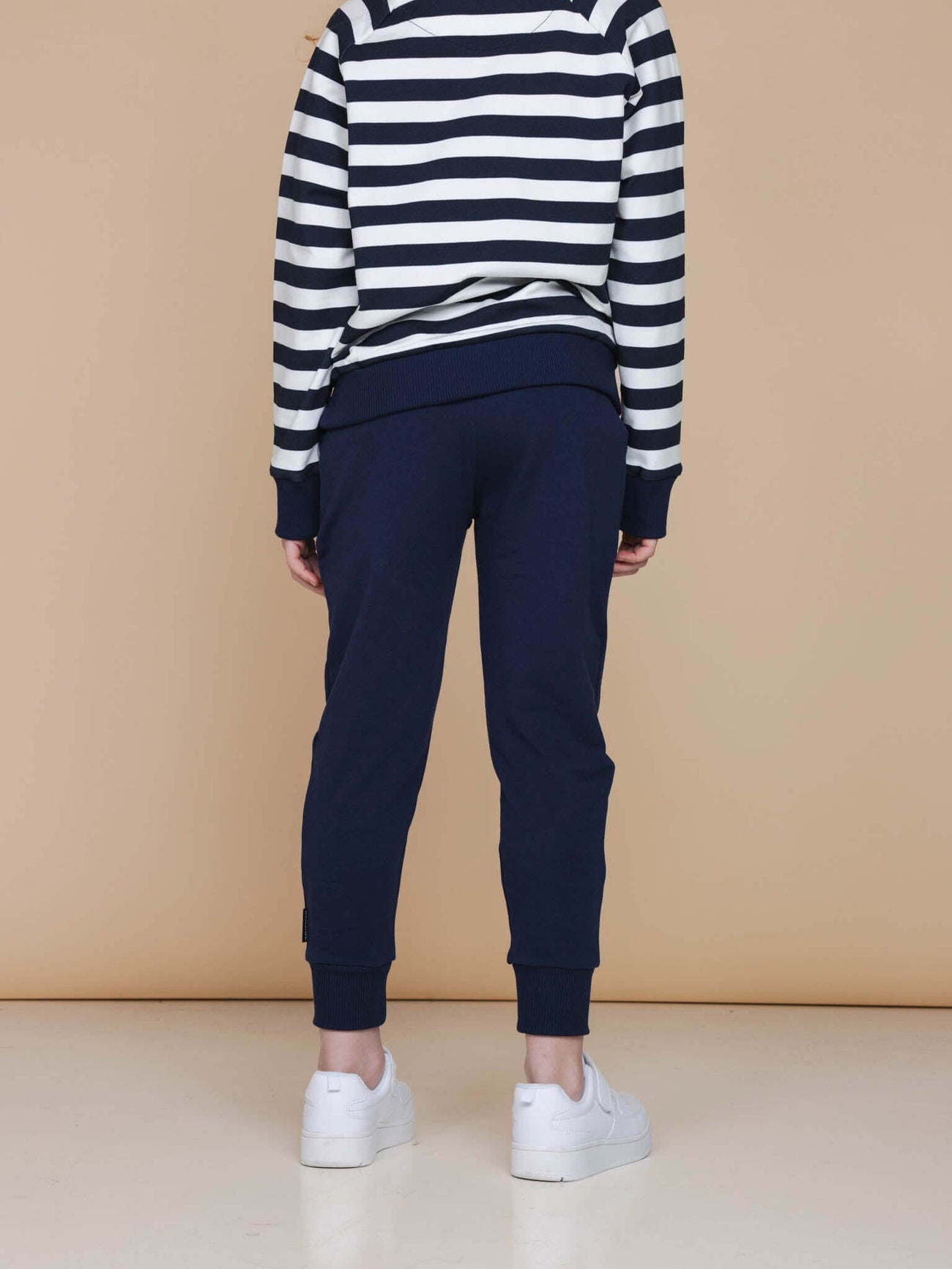 Kind in SNURK Navy Broek Kids, donkerblauwe joggingbroek met gestreept shirt en witte sneakers.