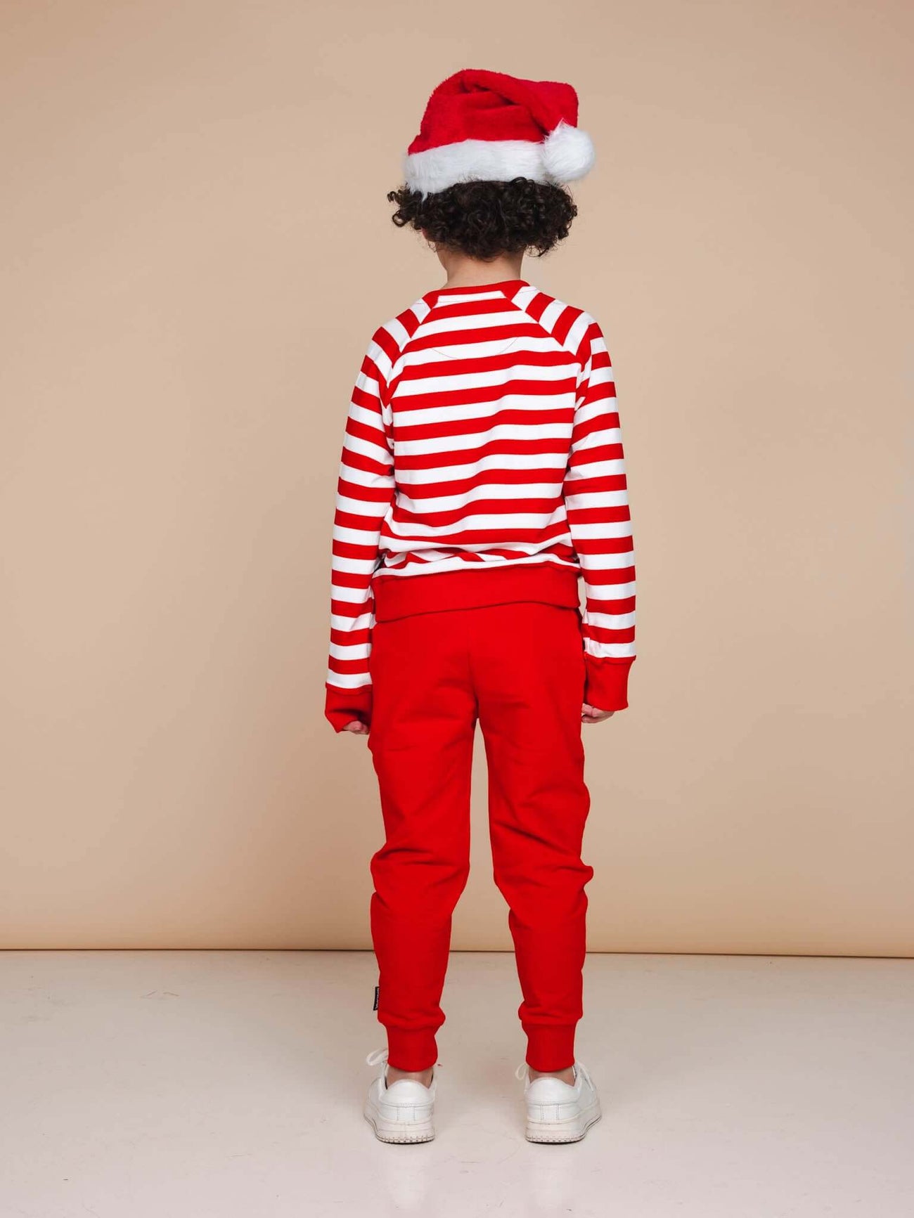 Een kind in een gestreepte trui en SNURK Red Broek Kids met een kerstmanmuts, perfect voor de feestdagen.
