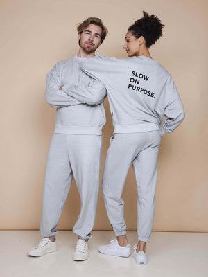SNURK Slow Grey Sweater Unisex worn by a man and woman, featuring 'SLOW ON PURPOSE' text on the back.